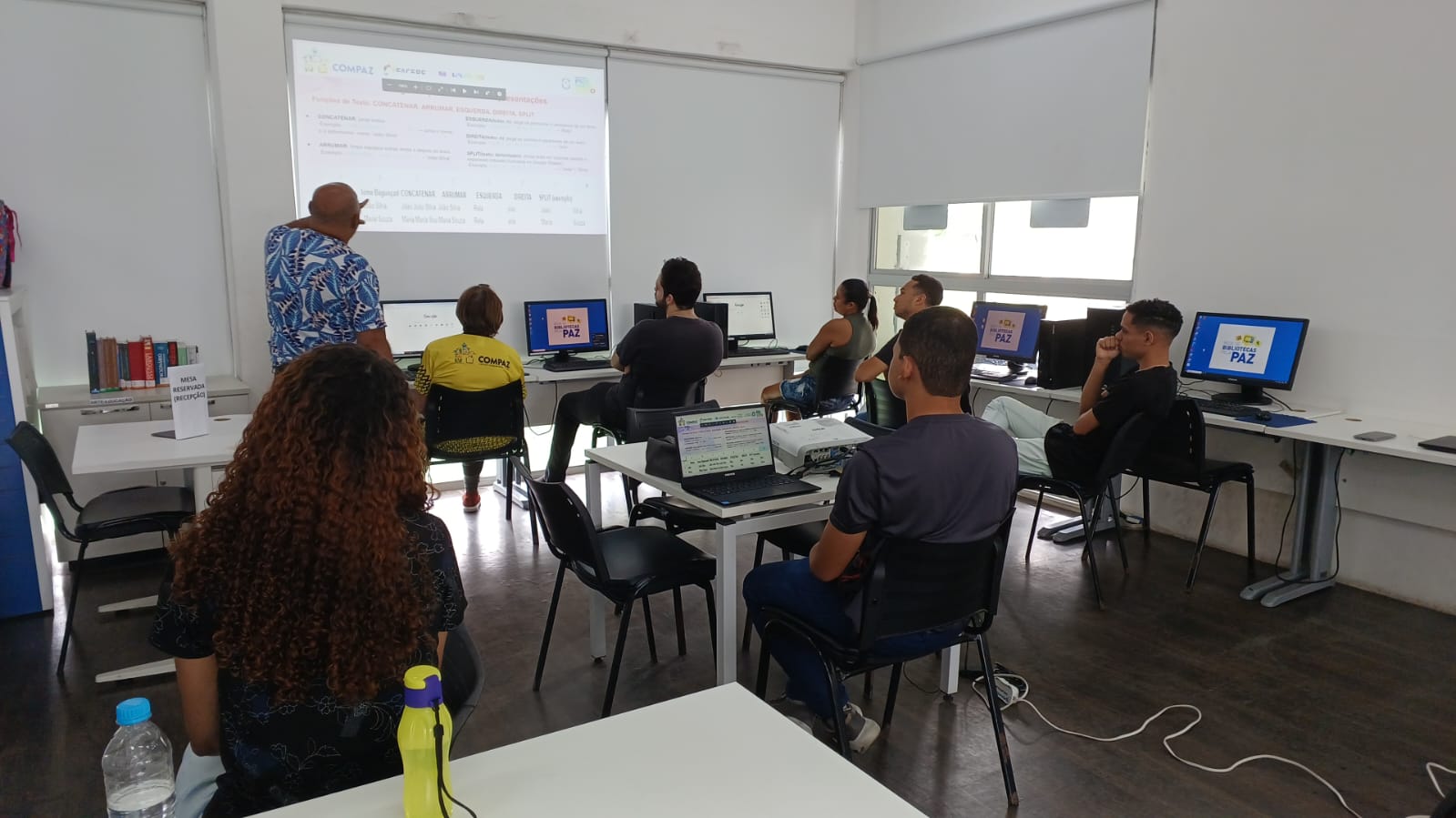 Bolsista do projeto ministra uma aula na biblioteca do COMPAZ. Ele está em pé, apontando para um slide em um projetor, enquanto os participantes assistem em seus computadores.