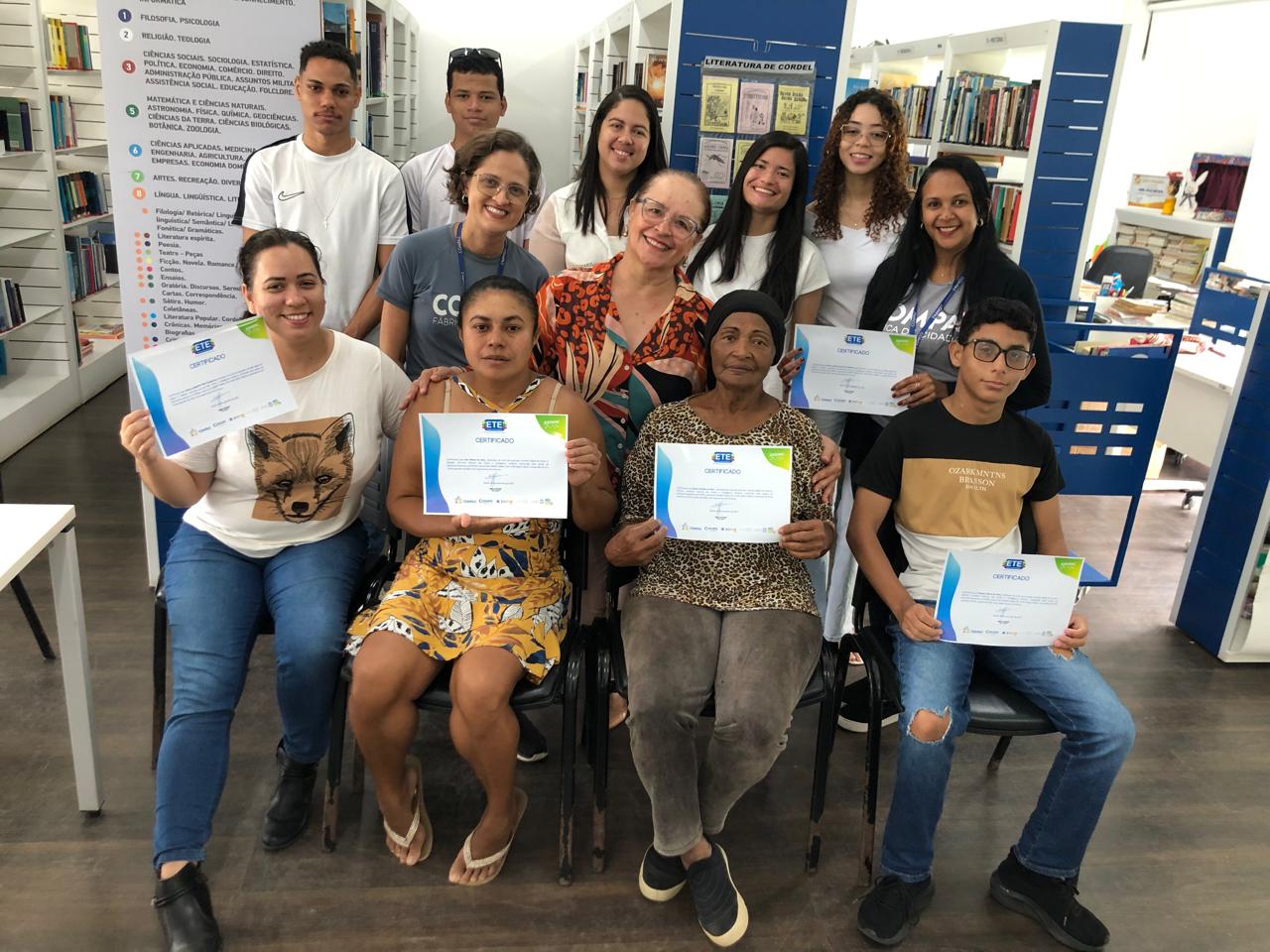 Alunos sentados à frente, sorrindo e exibindo seus certificados. Atrás, os bolsistas, a coordenadora e a bibliotecária.